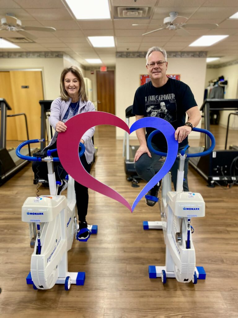 Two people smile while sitting on stationary bikes during cardiac rehab, each holding one side of a large heart-shaped cutout, with gym equipment and wooden floors visible in the background.