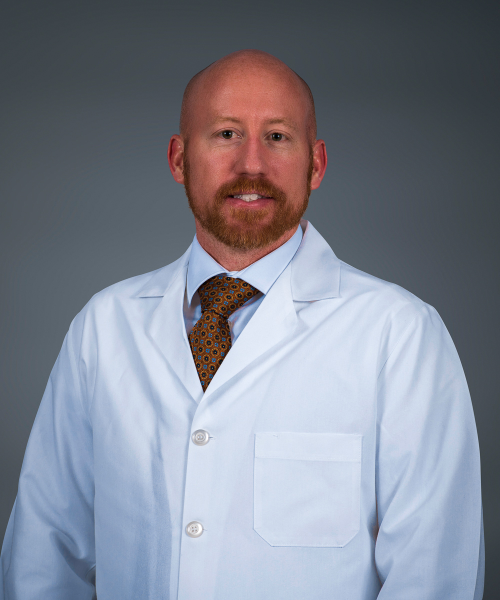 A man with a reddish beard and bald head wearing a white lab coat, dress shirt, and patterned tie stands in front of a plain dark background, looking at the camera.