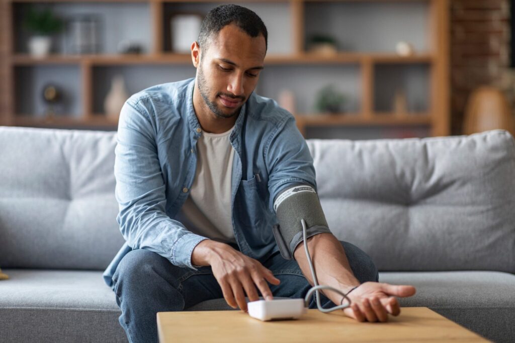 A man sitting on a sofa uses a digital blood pressure monitor, with the cuff on his arm and his finger pressing a button on the device, in a home living room setting.
