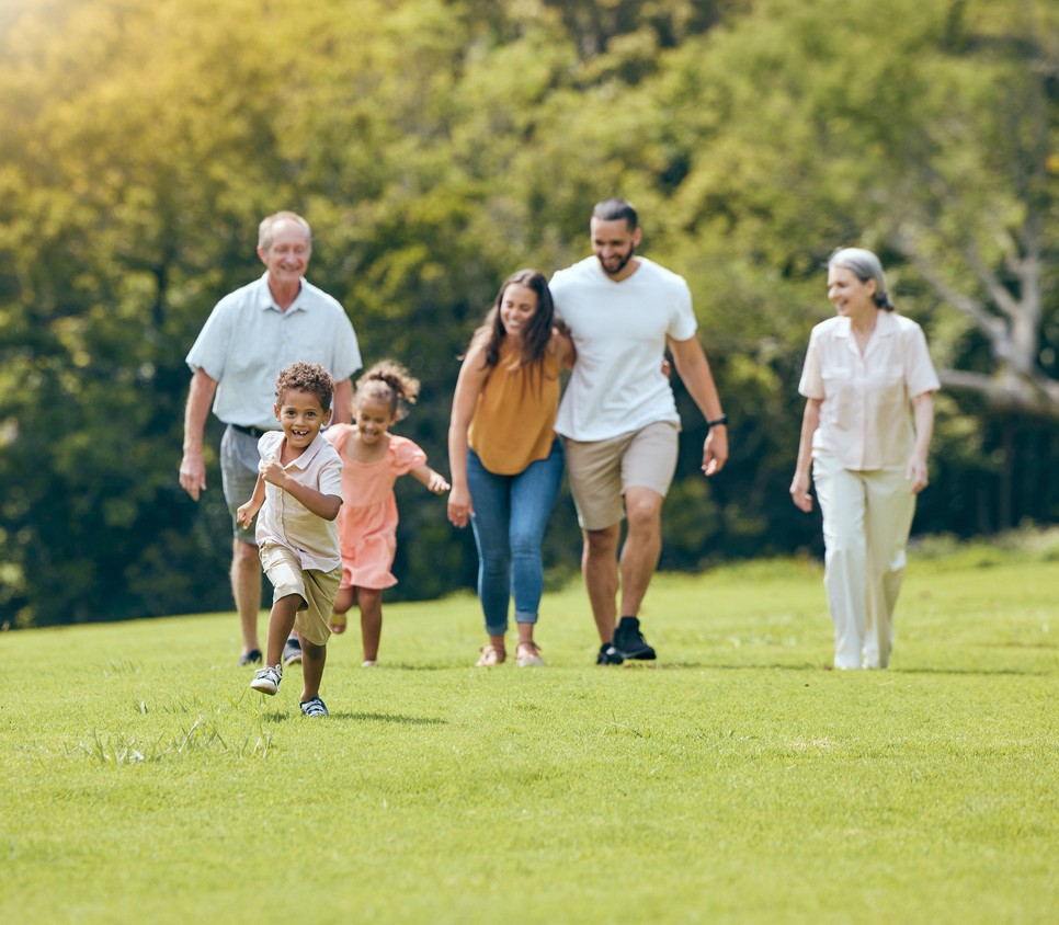 A young boy runs on grass in a park while a young girl and four adults, likely his family, including seniors, walk behind him, all smiling and enjoying a sunny day outdoors. Trees fill the background.