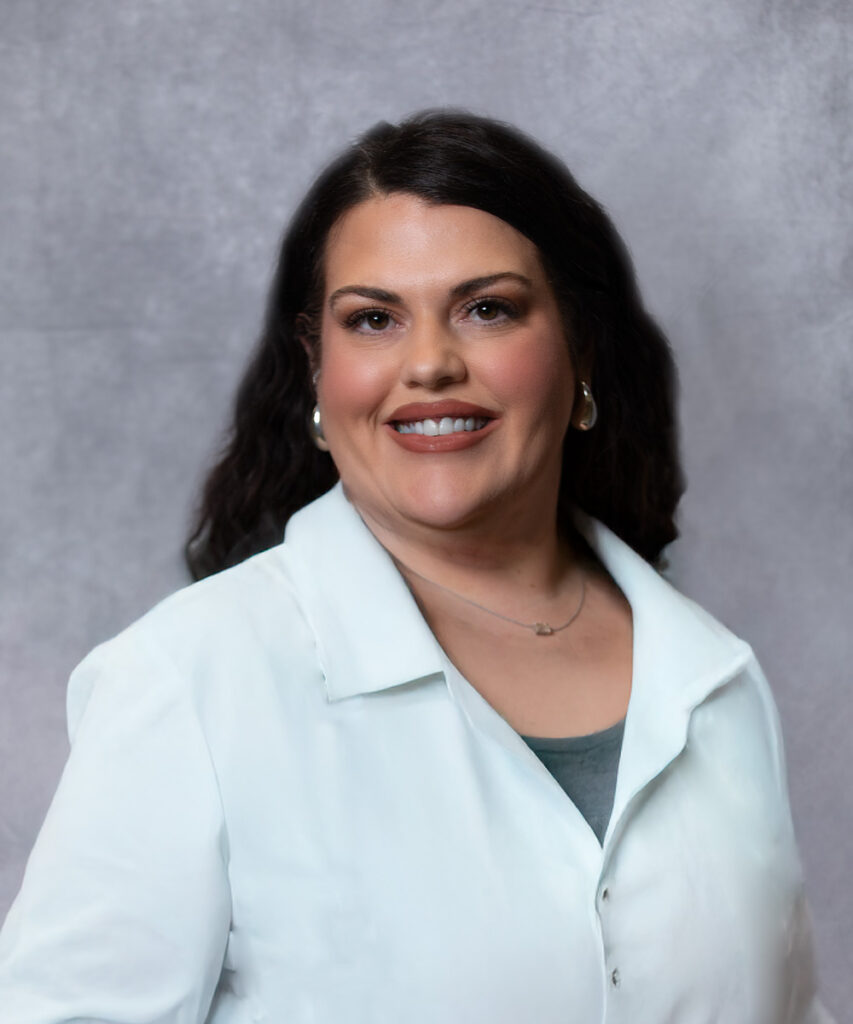 A woman with long dark hair wearing a white lab coat and a gray top smiles in front of a gray, textured background. She has hoop earrings and a delicate necklace.