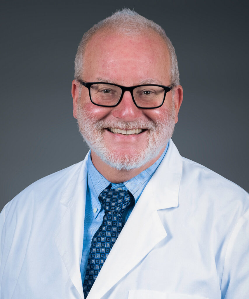 A smiling man with short white hair, a beard, and glasses is wearing a white lab coat, a light blue shirt, and a patterned tie, posing against a plain dark background.