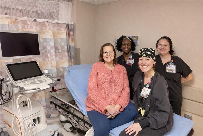 A woman in a pink sweater sits on a hospital bed, smiling, with three nurses in black scrubs standing beside her in a medical examination room with equipment and a monitor.