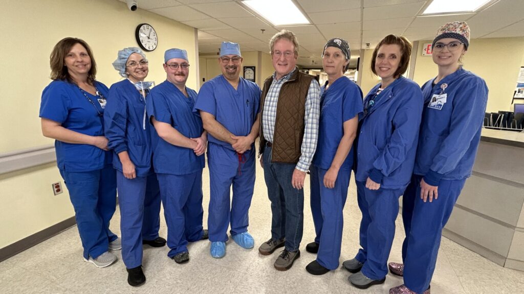 A group of eight people, including medical staff in blue scrubs and two people in casual clothing, stand together and smile in a hospital hallway. A clock and medical equipment are visible in the background.