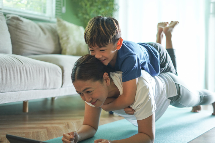 A woman does a plank exercise on a yoga mat while smiling, with a young boy lying on her back and hugging her. They are indoors with a couch and sunlight coming through a window in the background.