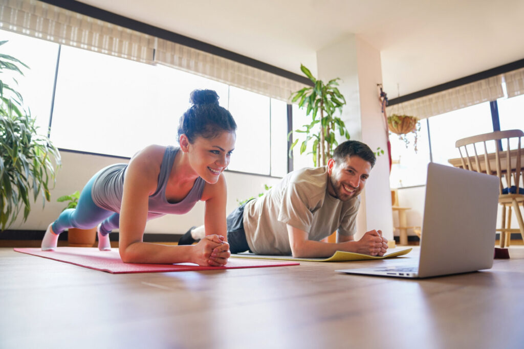 A woman and a man exercise together indoors, smiling while holding a plank position on yoga mats, following a workout on a laptop. Sunlight streams through large windows and plants decorate the room.