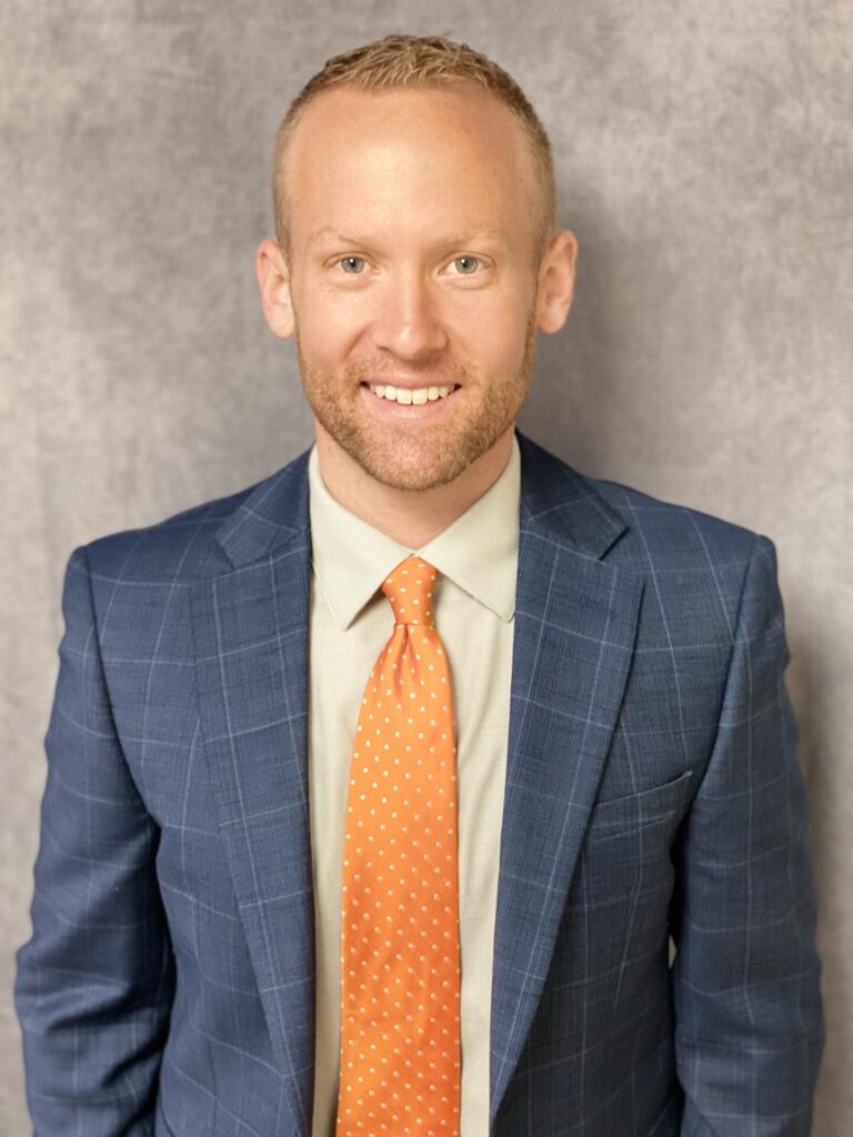 A smiling man with short blond hair wears a blue plaid suit, light shirt, and orange polka dot tie, standing in front of a light gray textured background.