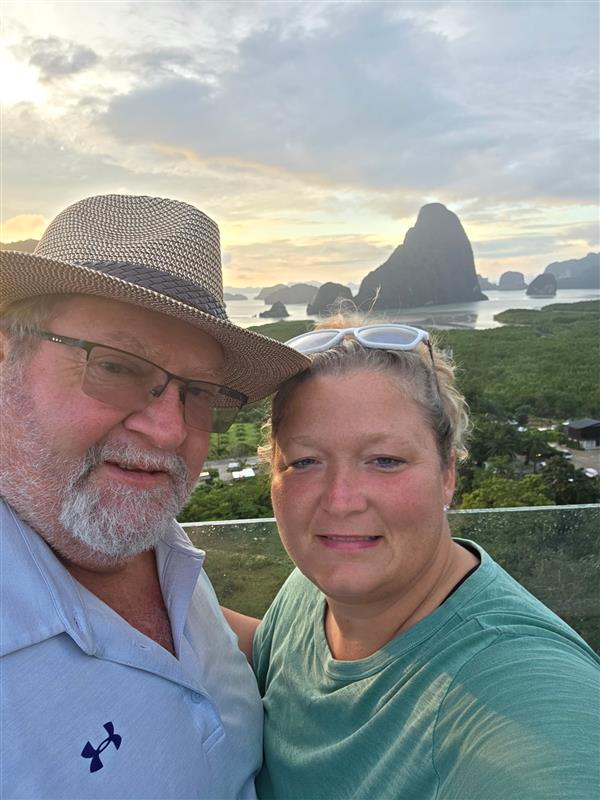 A man in a hat and glasses and a woman with sunglasses on her head smile for a selfie with lush greenery, water, and large limestone cliffs in the background at sunset.