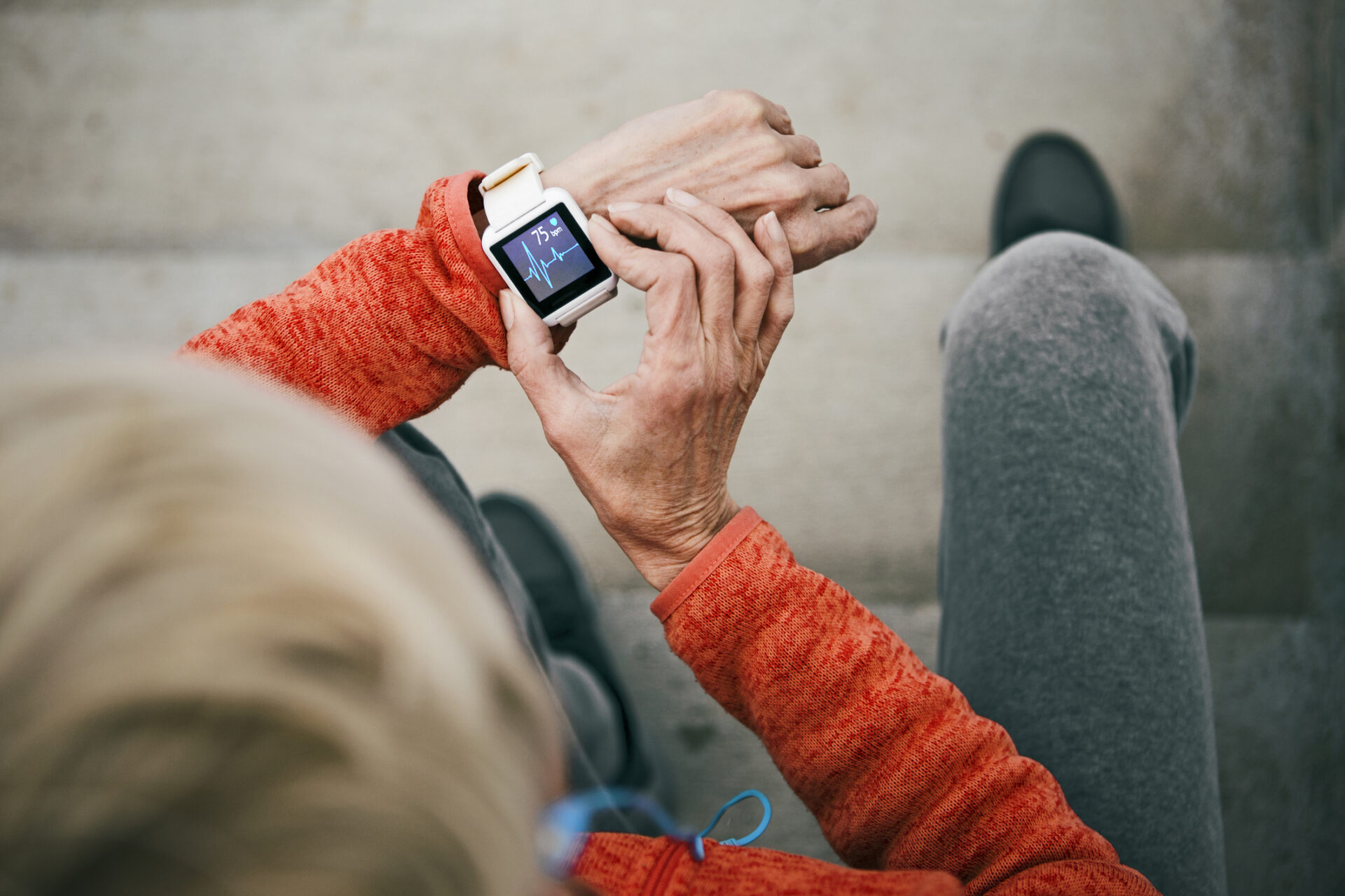 Person in a red jacket sitting on steps, checking their smartwatch, which displays a heart rate reading. The scene is viewed from above, focusing on the watch and the persons hands.