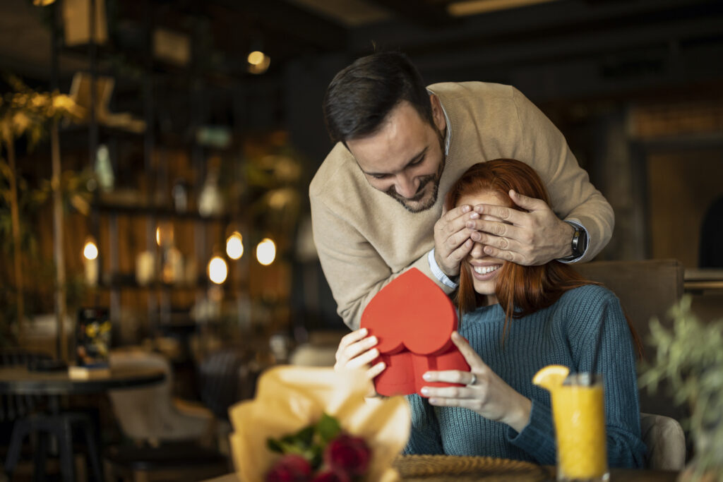 A man playfully covers a womans eyes as she smiles and holds a red heart-shaped gift box in a cozy restaurant. A bouquet of flowers and a drink are on the table in front of her.