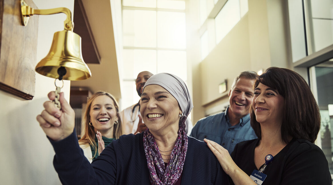 A smiling woman wearing a headscarf rings a bell on the wall, surrounded by cheering supporters and a medical professional in a bright hospital setting—a moment of hope made possible by caring teams and rewarding nursing jobs at Covenant Health.