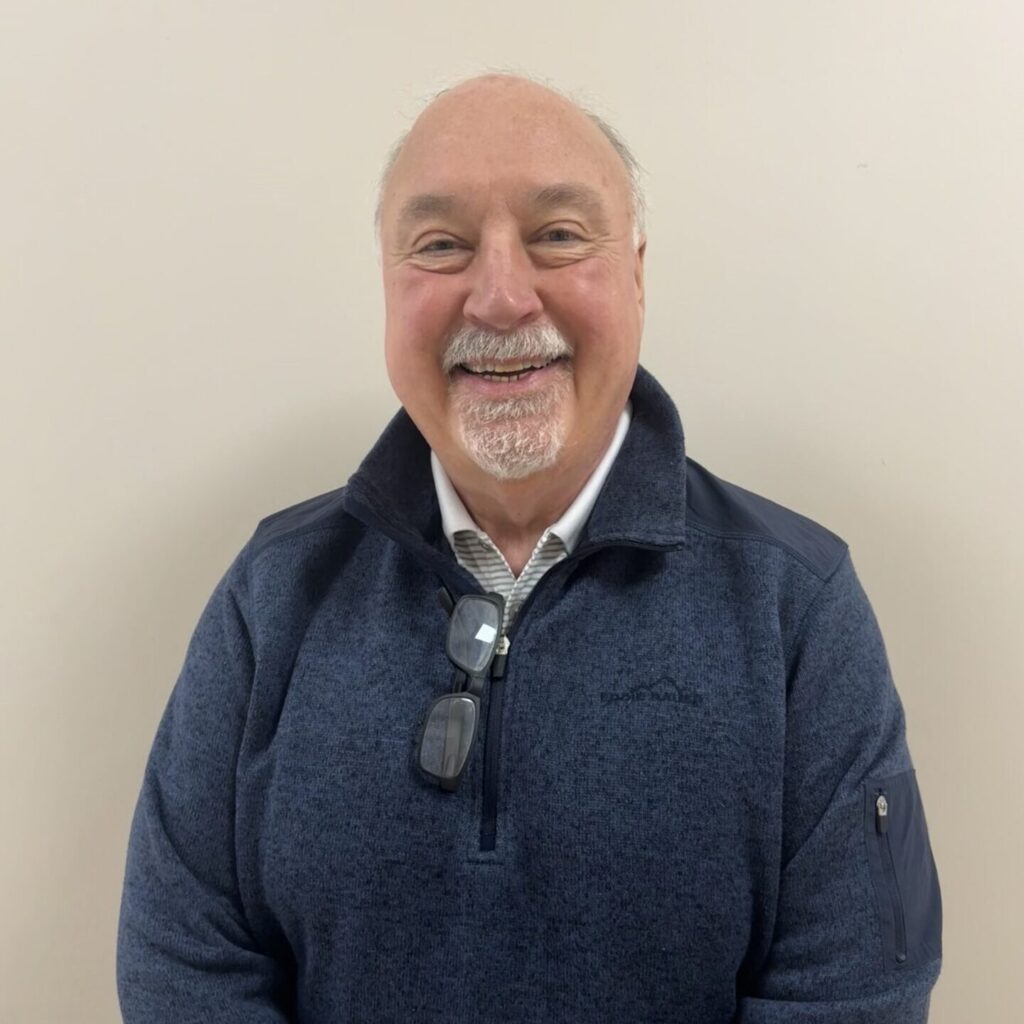 A smiling older man with short white hair and a mustache, wearing a dark blue pullover with glasses hanging from the collar, stands against a plain light-colored wall.