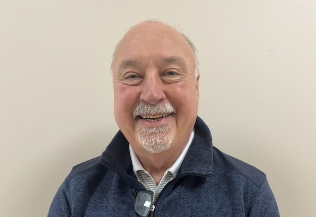 A smiling older man with short white hair and a mustache, wearing a dark blue pullover with glasses hanging from the collar, stands against a plain light-colored wall.