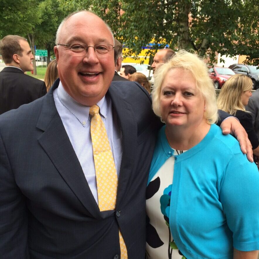 A smiling man in a suit and yellow tie stands with his arm around a woman in a blue cardigan and floral dress. They are outdoors among a group of people, with trees and greenery in the background.