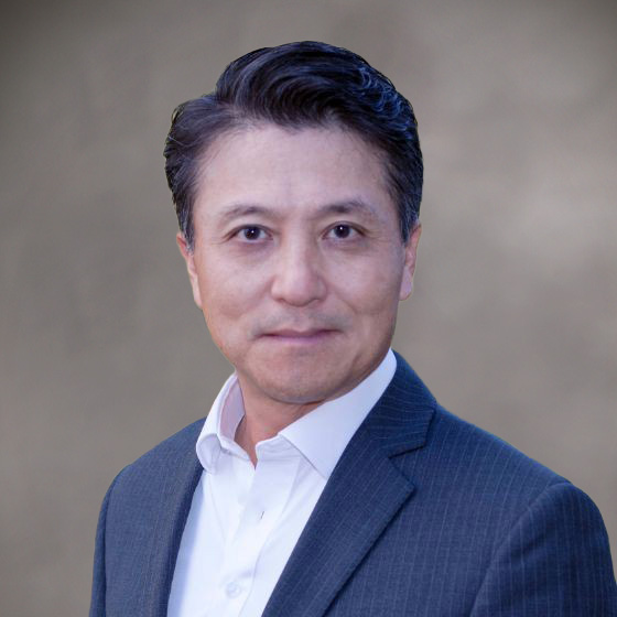 Robert Boos, a man with short, dark hair, wears a white collared shirt and a dark pinstripe suit jacket as he poses against a plain, blurred beige background.