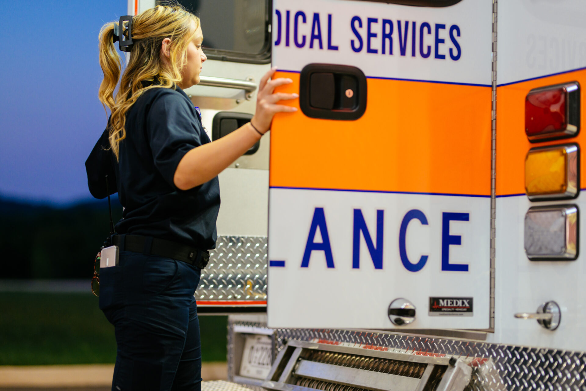 A paramedic in uniform stands beside an open ambulance door, holding it with one hand, preparing to enter the vehicle. The ambulance has bright orange and blue markings.