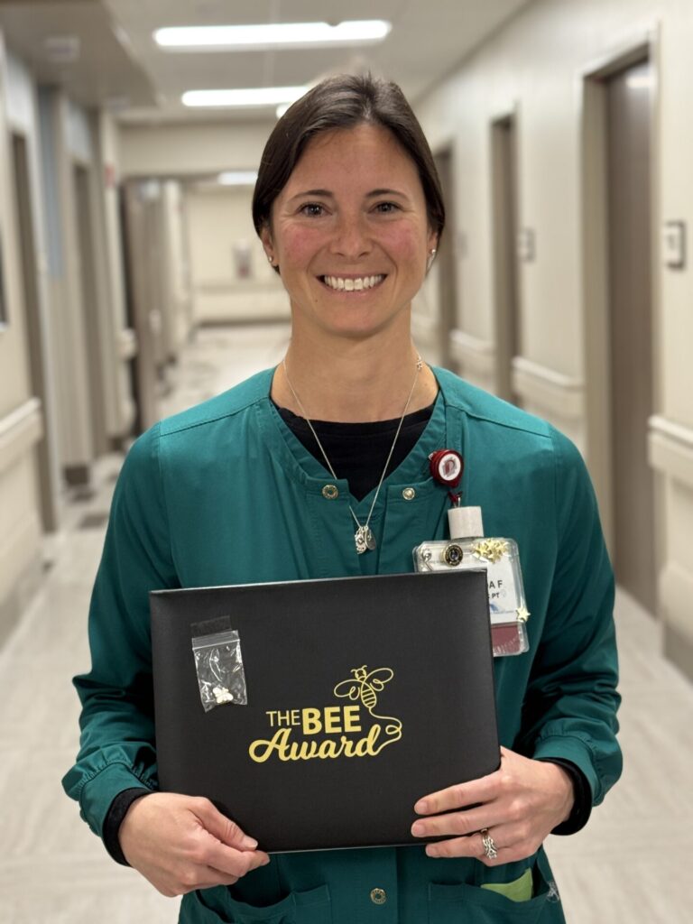A woman in green scrubs stands in a hallway, smiling and holding a black folder labeled The BEE Award in gold letters. She wears a work badge identifying her as Ahniah Ray and appears to be in a professional healthcare setting.