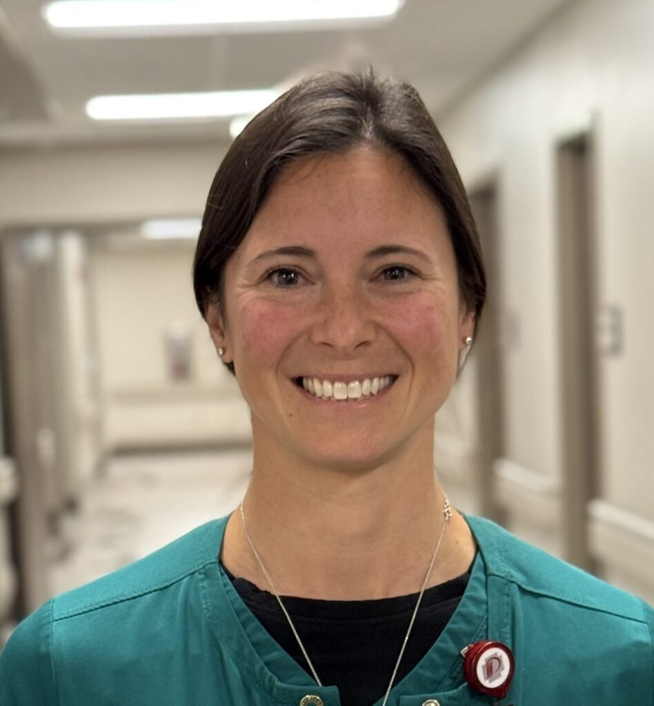 A woman with short brown hair and a green top smiles in a brightly lit hallway, possibly in a hospital or clinic, proudly wearing her badge and necklace as the recipient of the Ahniah Ray BEE Award.