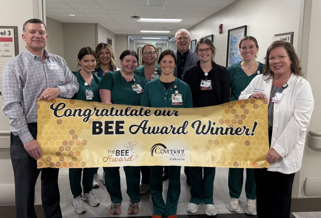 A group of medical staff in green and white uniforms stand in a hallway, smiling and holding a yellow banner that says Congratulate BEE Award Winner! with honeycomb patterns.