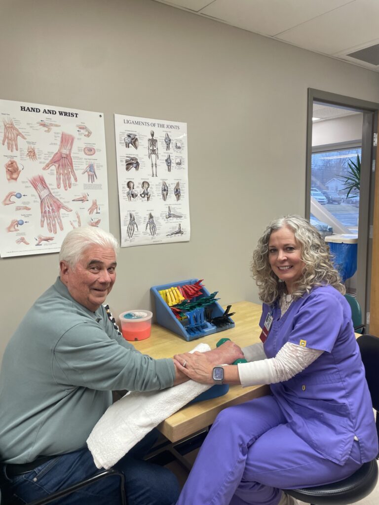 An older man sits at a table with a smiling healthcare worker in purple scrubs, who is treating his hand. Medical diagrams of the hand and wrist are on the wall behind them.