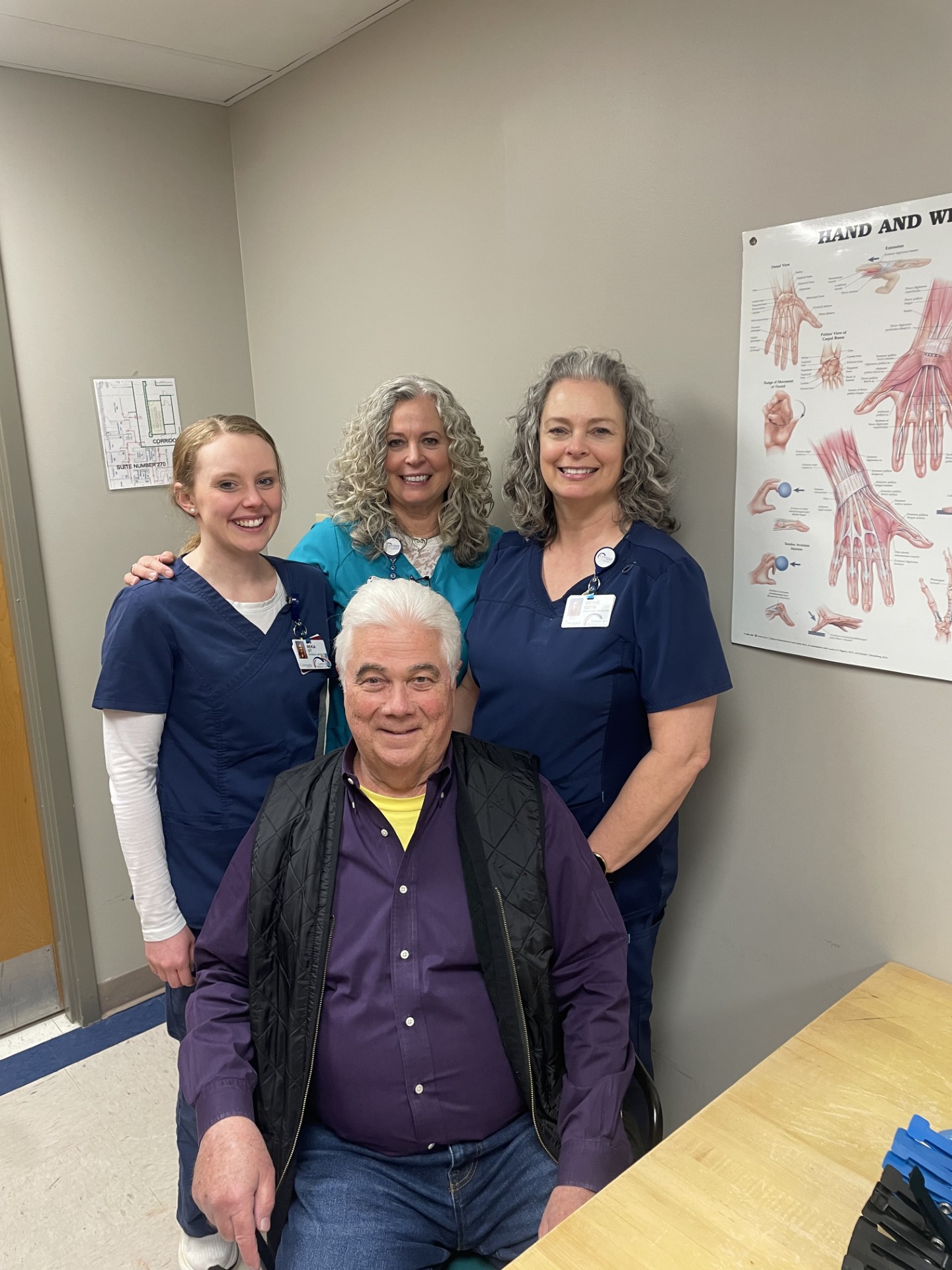 Three women in medical scrubs stand smiling behind an older man sitting in a chair. They appear to be in a medical office, with a poster of hands and wrists anatomy visible on the wall.