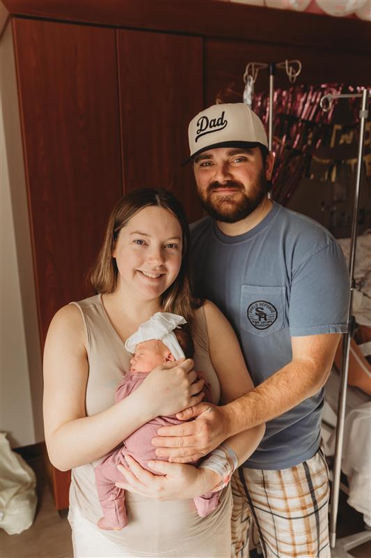 A smiling woman holds a newborn baby wrapped in a pink blanket, while a man wearing a Dad hat stands beside them with his arm around her, in a warmly lit room.