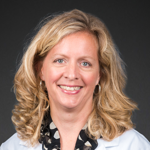A woman with wavy blonde hair, wearing a white coat and a black blouse with white spots, smiles at the camera against a plain dark background.