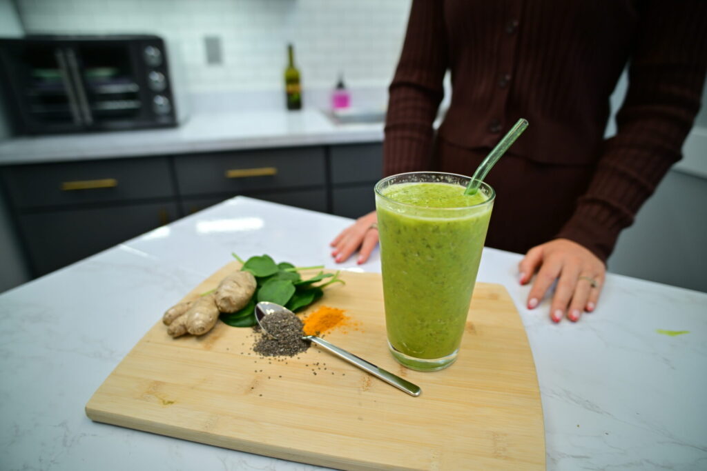 A person stands behind a cutting board with ginger, spinach, turmeric, chia seeds, and a spoon, next to a glass of green smoothie with a straw on a kitchen counter.