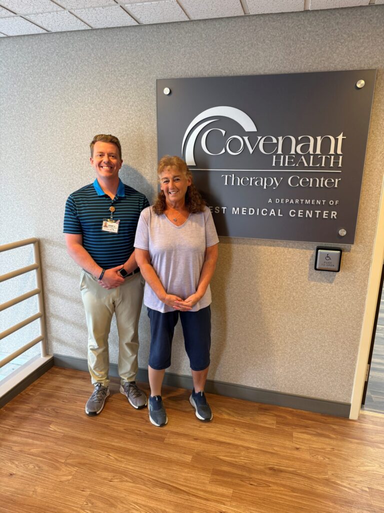 A man and a woman stand smiling in front of a wall sign that reads Covenant Health Therapy Center, A Department of West Medical Center inside a medical facility.