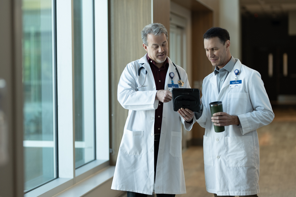 Two male doctors in white lab coats stand by a window, looking at a tablet together. One holds a coffee tumbler. They appear to be discussing nursing jobs at Covenant Health in a brightly lit hospital hallway.
