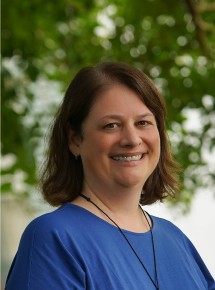 A woman with shoulder-length brown hair, wearing a blue top and a necklace, smiles outdoors with green trees blurred in the background.