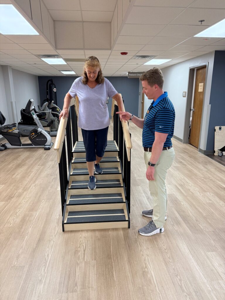 A woman practices walking down a set of therapy stairs with handrails, guided by a physical therapist in a rehabilitation center with exercise equipment in the background.