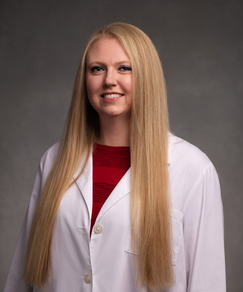 A woman with long, straight blonde hair wearing a white lab coat over a red top, smiling and standing in front of a neutral gray background.