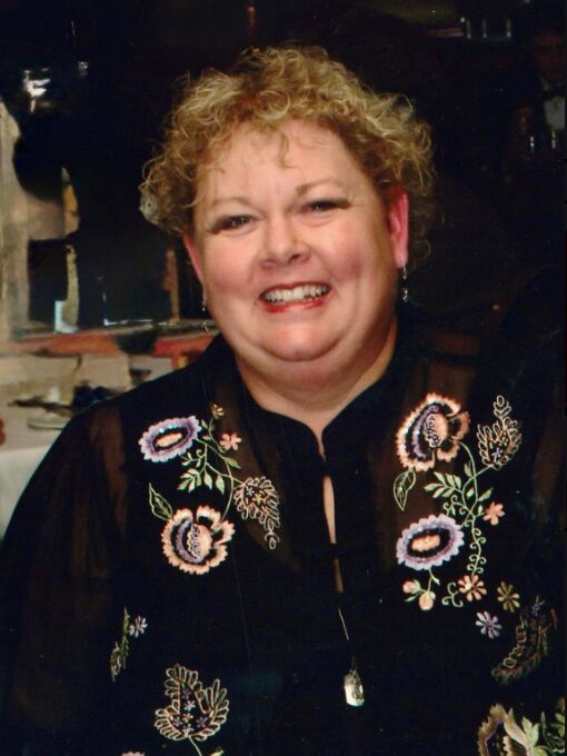 A smiling woman with curly blonde hair wears a black top with colorful floral embroidery and a pendant necklace, standing indoors with a blurred background.