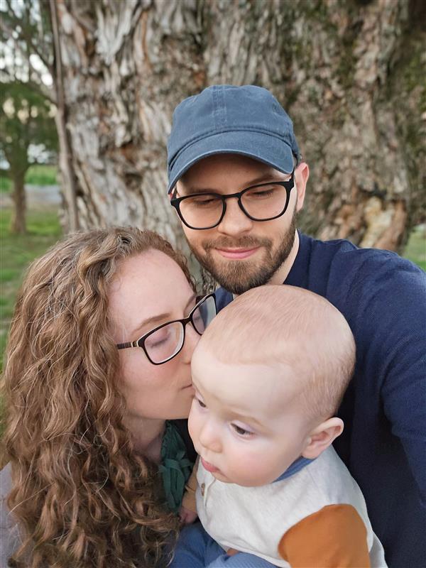 A woman with curly hair and glasses kisses a baby on the cheek while a man in a blue cap and glasses smiles beside them. They are outdoors near a tree.