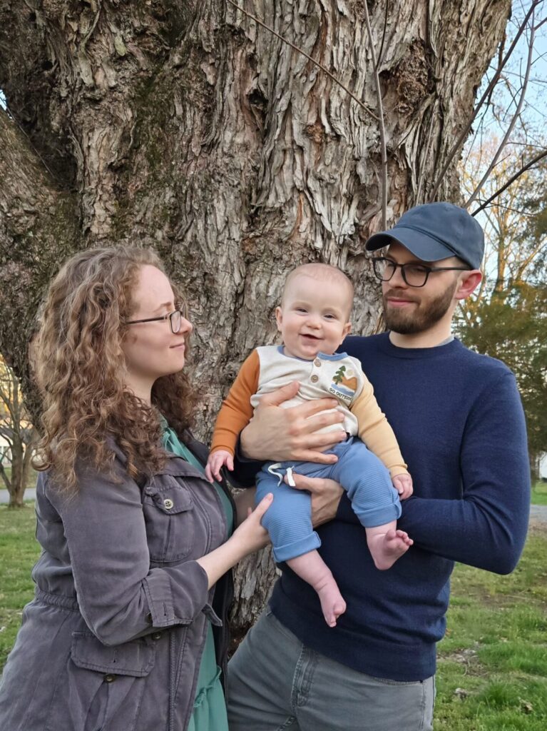 A woman and a man stand in front of a large tree, holding a smiling baby between them. The woman has curly hair and glasses, and the man wears a cap and glasses. All look content outdoors in a park setting.