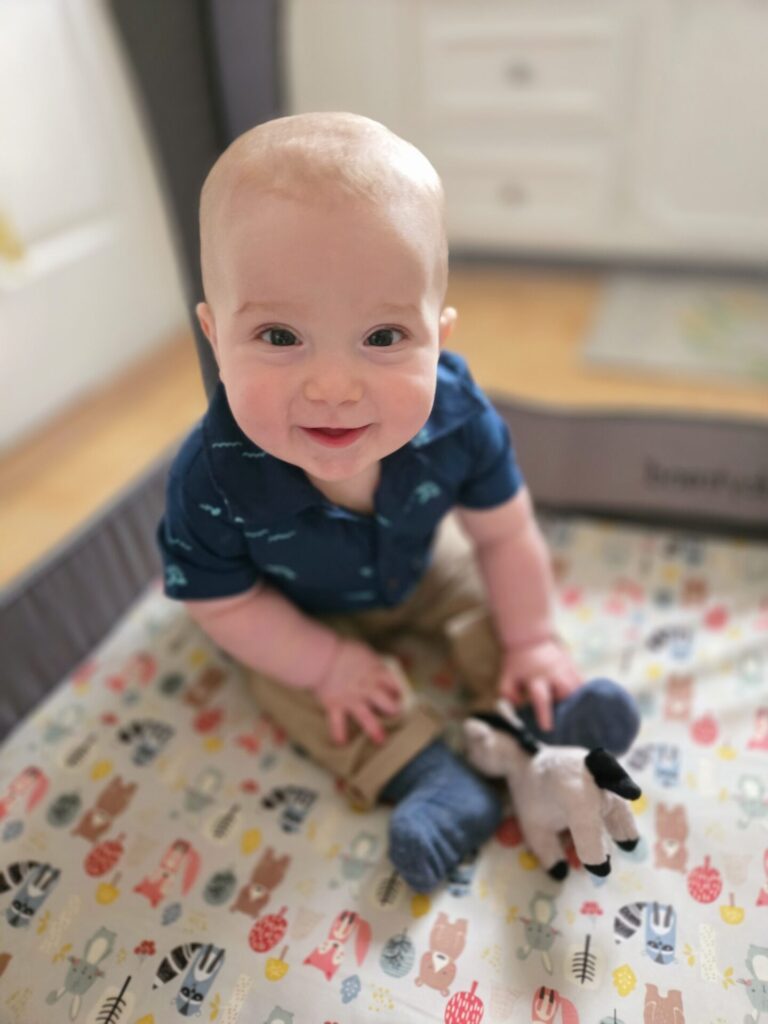 A smiling baby with light skin and light hair sits in a playpen on a colorful animal-patterned mat, wearing a navy blue shirt and khaki pants, with a plush toy and socks nearby.