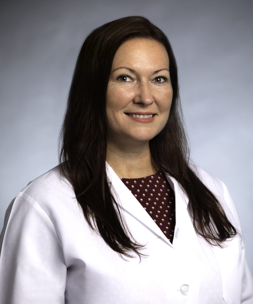 A woman with long brown hair, wearing a white lab coat over a maroon blouse with white polka dots, smiles at the camera against a light gray background.