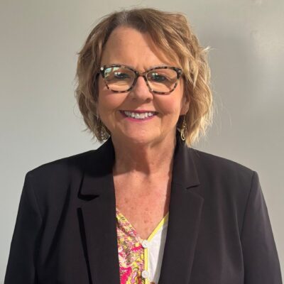 Smiling woman with short, wavy blonde hair and glasses, wearing a black blazer over a colorful blouse, standing in front of a plain light-colored background.
