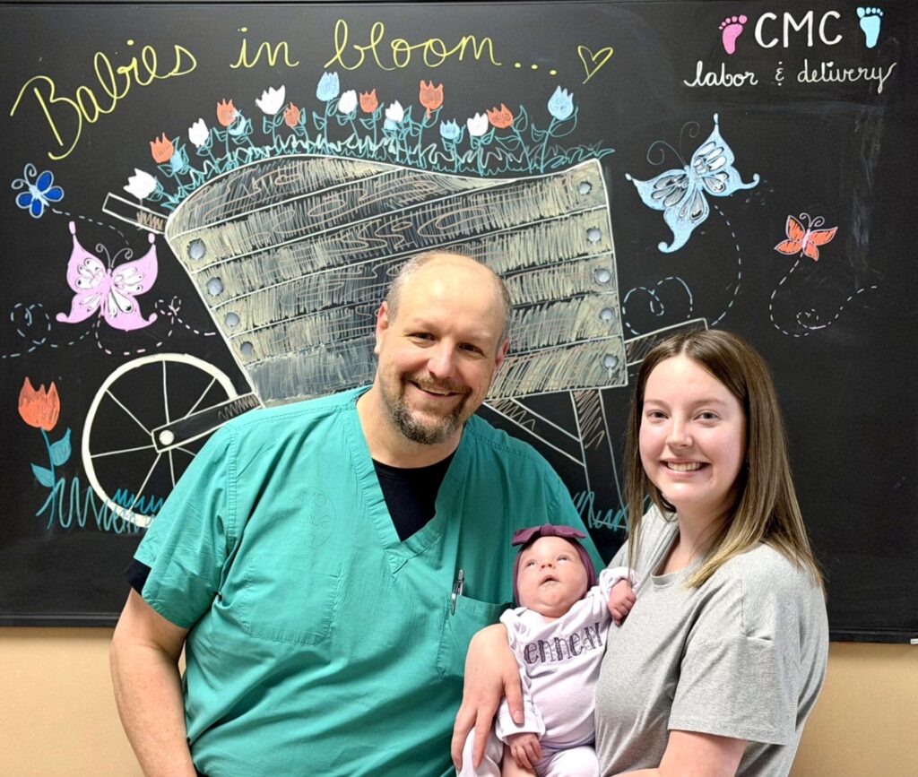 A man in green scrubs, a woman, and a baby pose and smile in front of a chalkboard with colorful drawings of flowers, butterflies, and the words Babies in bloom... CMC labor & delivery.