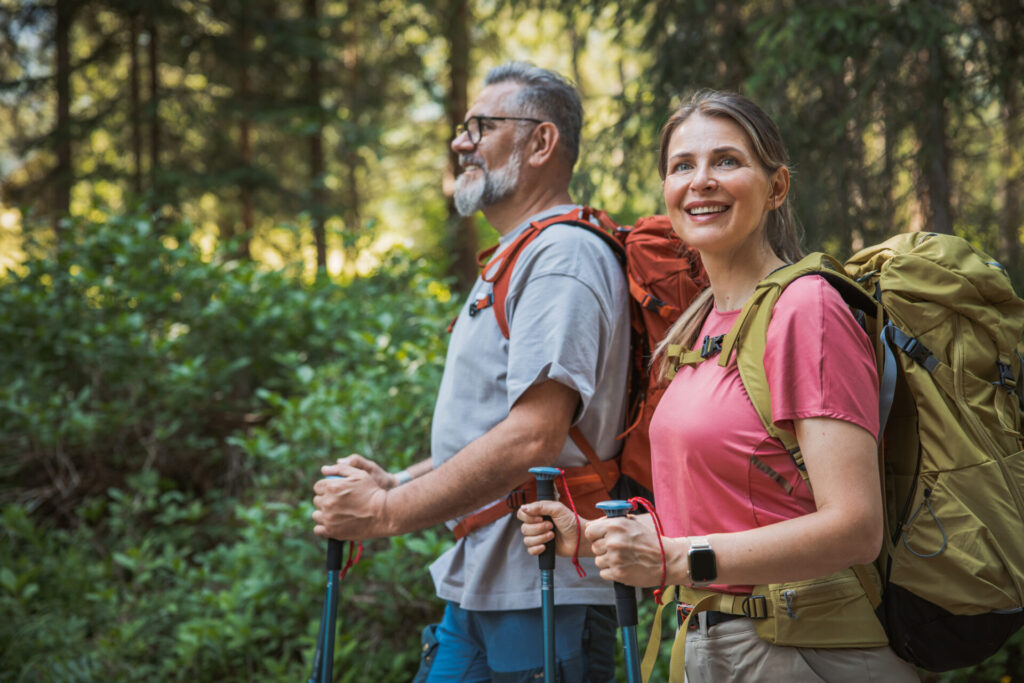 A man and woman with backpacks and trekking poles hike through a sunlit forest, smiling as they enjoy the outdoors. Surrounded by green foliage, they make cardiac rehab an active and peaceful journey in nature.