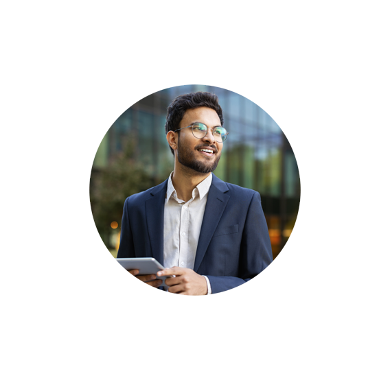 A man in a suit and glasses smiles while holding a tablet outdoors, with a modern glass building and greenery blurred in the background.
