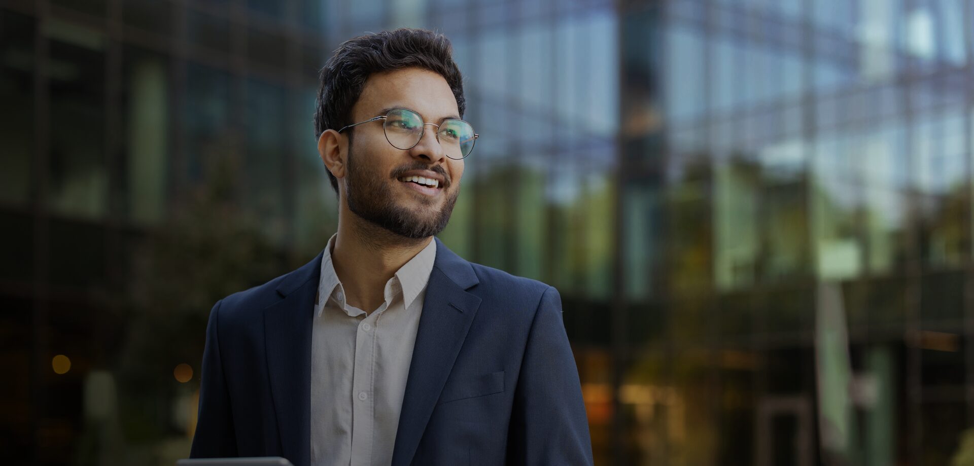 A man wearing glasses and a suit jacket smiles while standing outdoors in front of a modern glass office building.