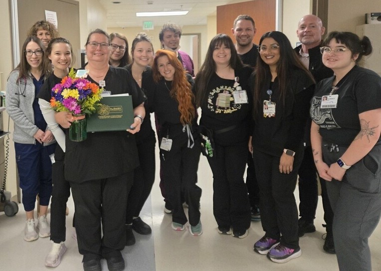 A group of healthcare workers stand together in a hospital hallway, smiling. Natasha Logan wins DAISY Award, holding a bouquet of colorful flowers and a certificate. Most are dressed in black scrubs, and they appear happy and celebratory.