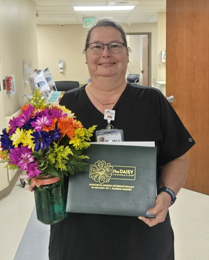 A nurse in black scrubs stands in a hallway, holding a colorful bouquet of flowers and a green DAISY Award folder, smiling at the camera as Natasha Logan wins DAISY Award.