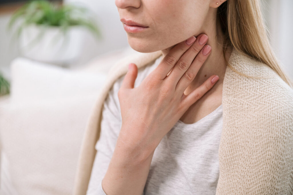 A woman touches her throat with her hand, appearing to check or soothe discomfort from a sore throat. She is sitting indoors with a light-colored blanket over her shoulders.