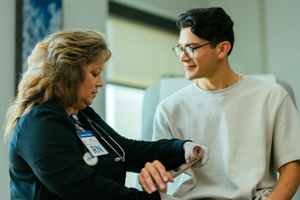 A nurse wearing a badge labeled RN checks the blood pressure of a smiling young man in a medical office. The nurse focuses on the man’s arm, which is wrapped in a blood pressure cuff.