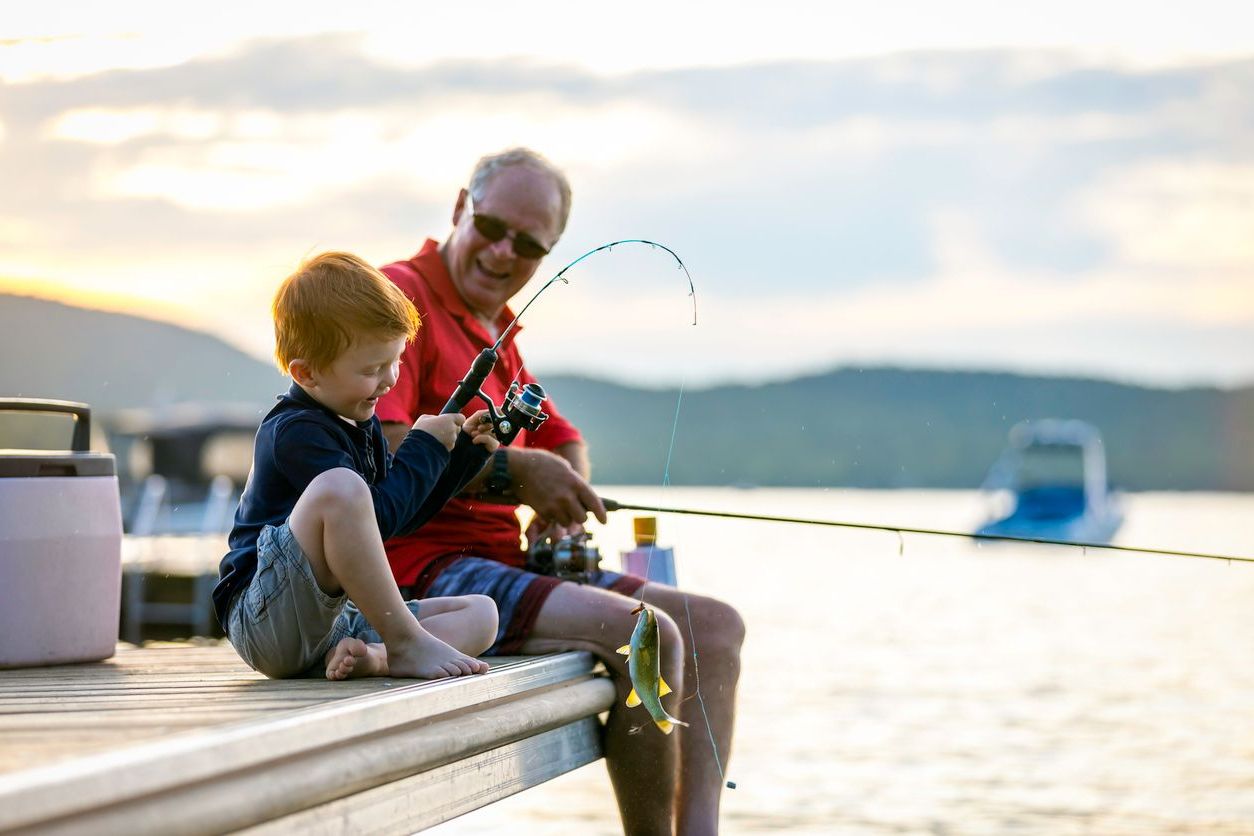 A young boy and an older man sit on a dock fishing together. The boy smiles, holding a fishing rod with a fish on the line, while the man looks on—both enjoying a sunny day by the water and sharing moments of covenant health and happiness.