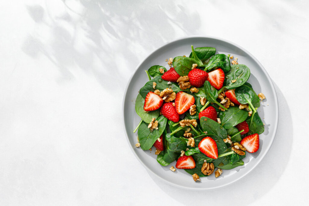 A white plate with a fresh salad of spinach leaves, halved strawberries, and walnuts on a white background with soft shadows.