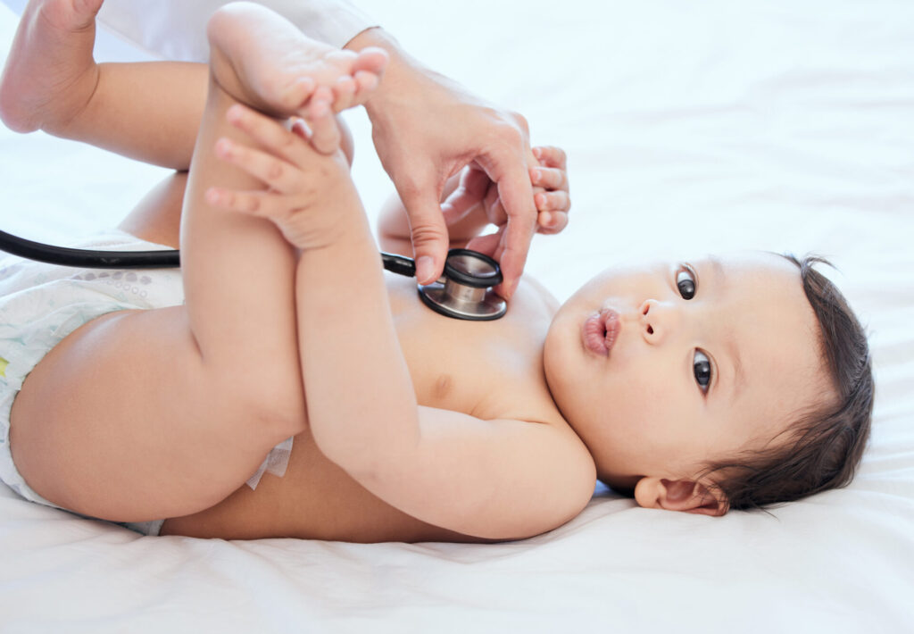 A baby lies on a white surface as a healthcare professional listens to its chest with a stethoscope&mdash;an important moment in learning How to Practice Safe Sleep for Babies. The baby holds its legs up and gazes curiously at the camera.